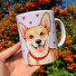 Prémium Corgi kutyás bögre, amelyen két különböző, kézzel rajzolt kutyagrafika látható: egyik oldalán közelről megfestett, részletgazdag Corgi arc, a másikon egész alakos, mosolygó kutya. A szívecskés háttér finom, akvarellhangulatú dekoráció. ☕🐾