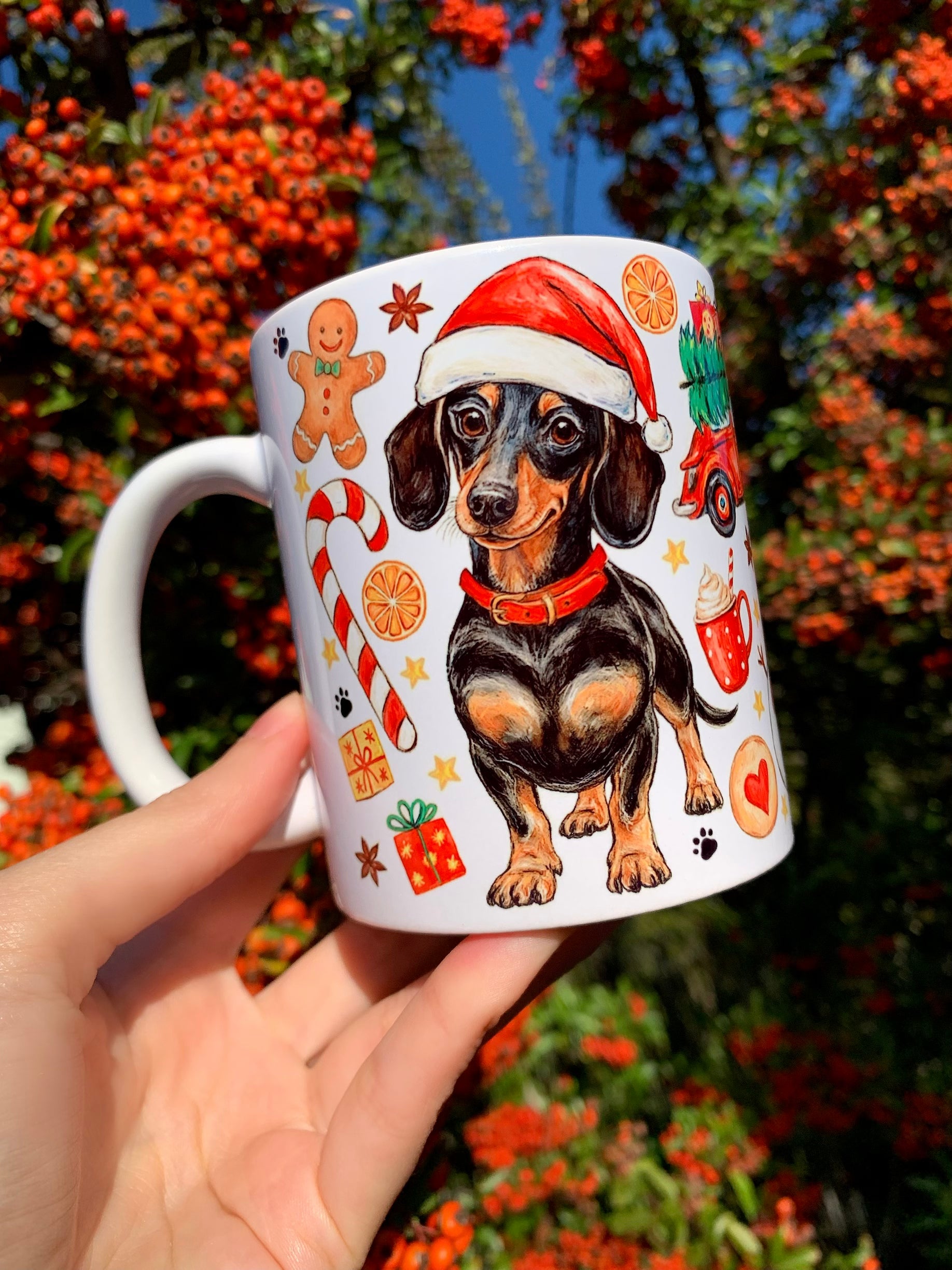 Karácsonyi hangulatú kerámia bögre művészien rajzolt tacskóval és piros nyakörvvel. A tacskót körülölelő piros–zöld dekorációk finom akvarellszínezést kaptak. Kézműves hatású illusztrációval készült.