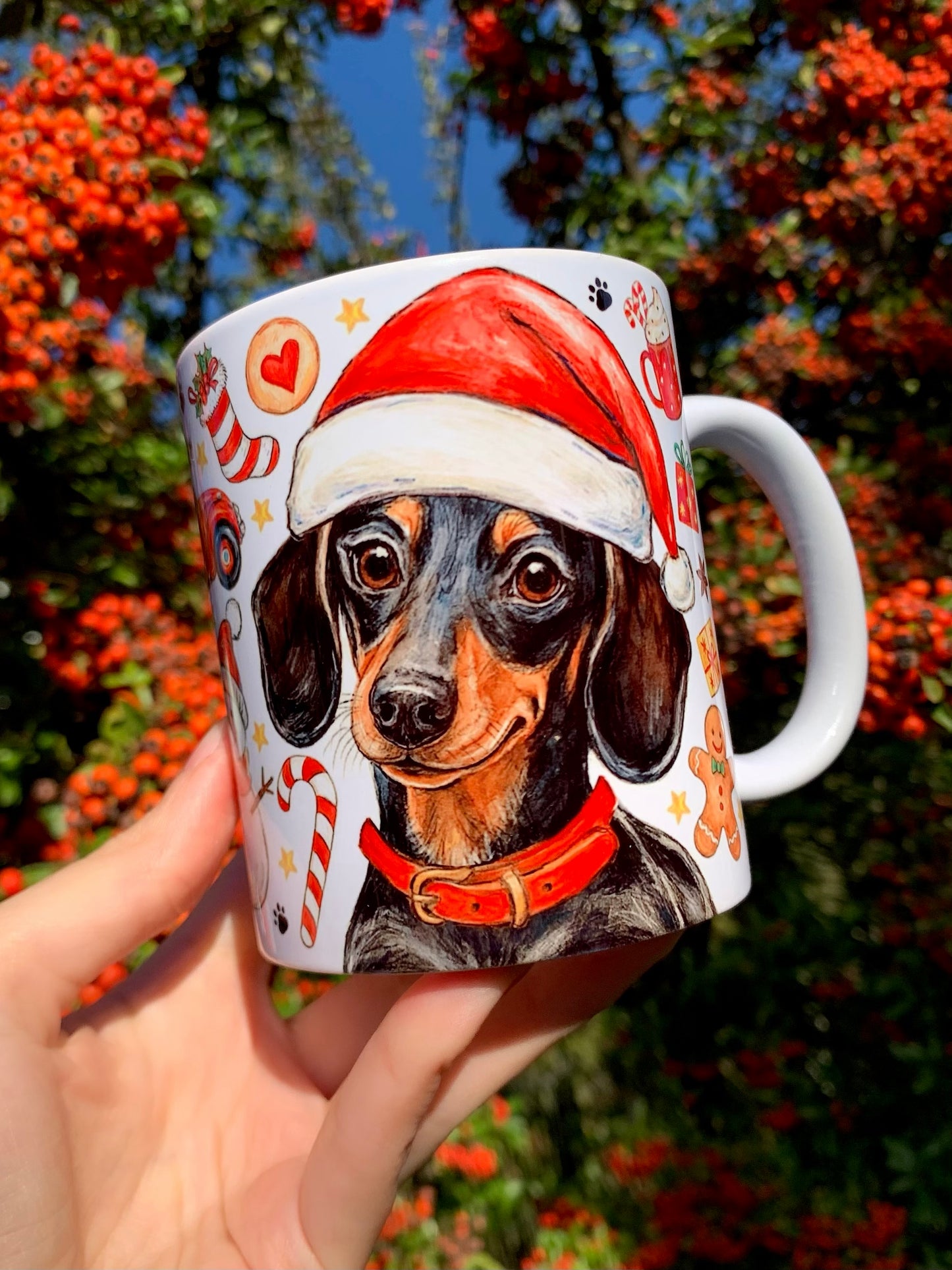 Bájos tacskós karácsonyi bögre kézműves, ceruzás–akvarelles illusztrációval. A minta gazdagon díszített ünnepi elemekkel: harangokkal, fenyőágakkal, mézeskaláccsal. Hangulatos, szerethető ünnepi design.