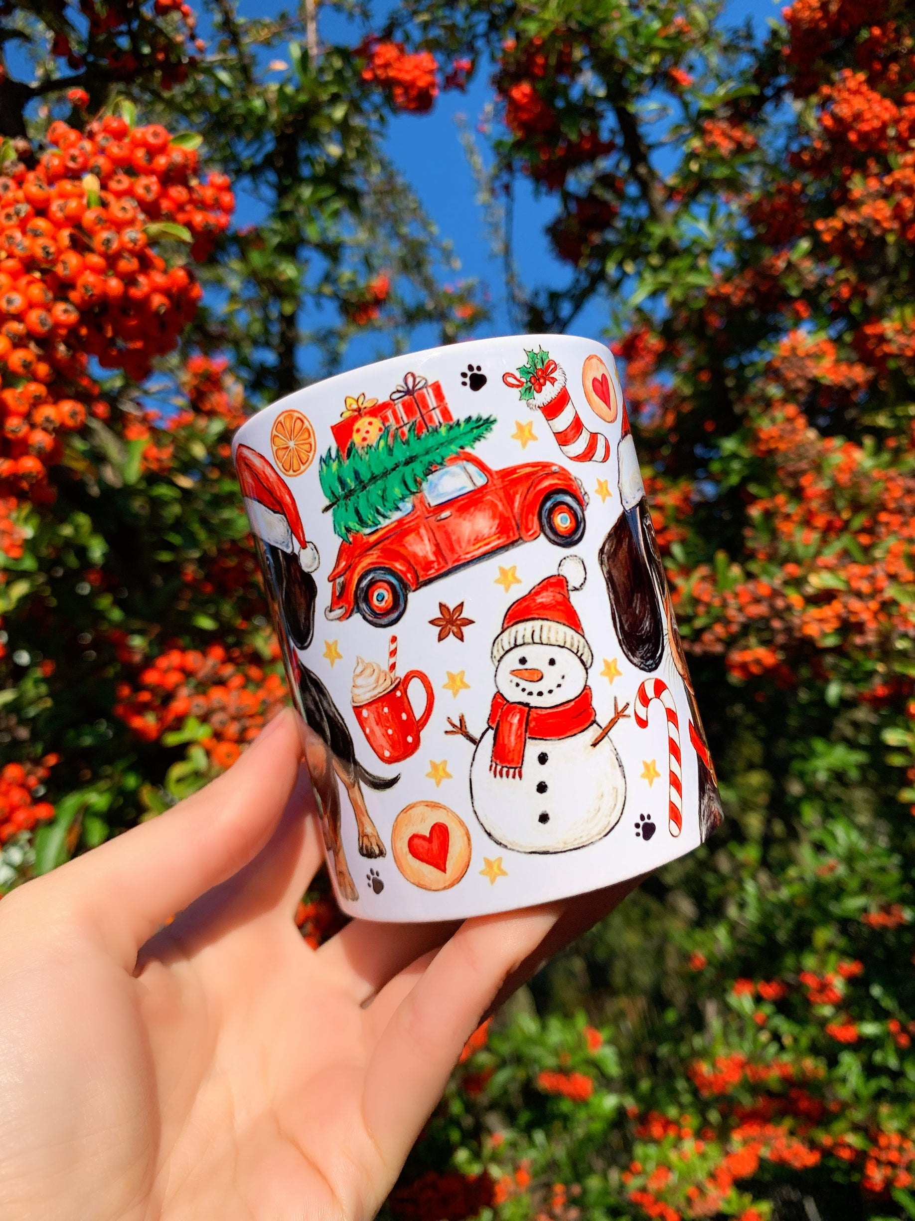 Kerámia bögre finoman megrajzolt, akvarelles tacskóval és piros nyakörvvel. A környező karácsonyi díszek – mézeskalács, cukorpálca, fenyőág – gazdag ünnepi keretet adnak. Kézműves, részletgazdag grafika.