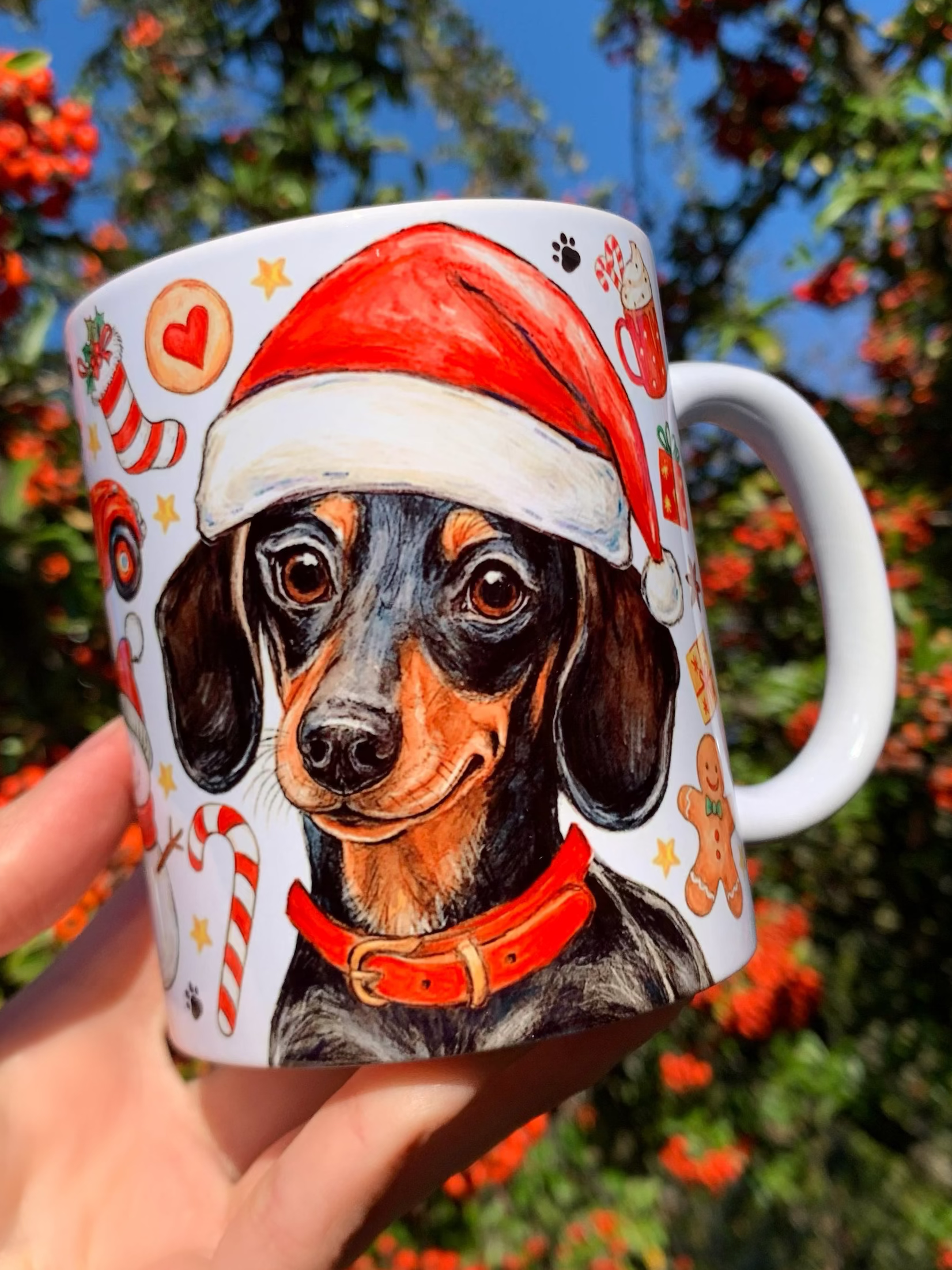 Prémium minőségű karácsonyi kerámia bögre kézzel rajzolt és akvarellel festett cser színű tacskóval, piros nyakörvvel és ünnepi motívumokkal. A részletgazdag kutyailusztráció köré mézeskalács figurák, piros–fehér cukorkák, arany harangok és zöld fenyőágak kerültek, finom akvarellárnyalatokkal. A kézműves stílusú minta meleg, ünnepi hangulatot teremt, tökéletes ajándékká téve a bögrét karácsonyra.
