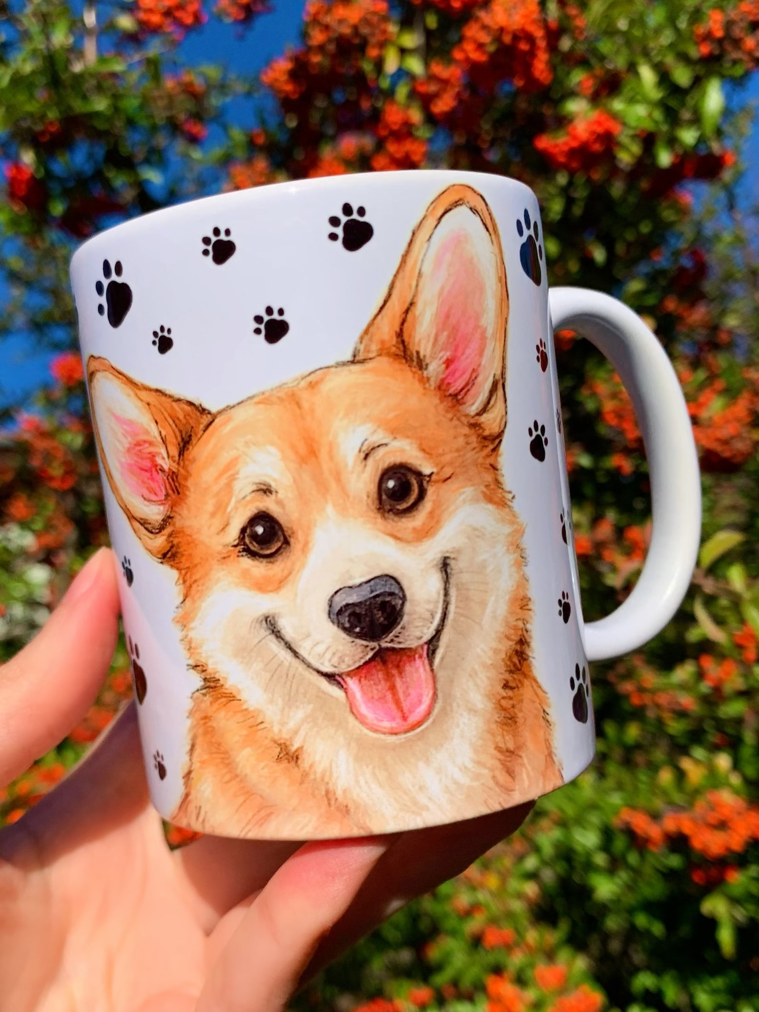 Kézzel festett Corgi kutyás kerámia bögre, Pusztai Juci részletgazdag illusztrációjával: mosolygó Corgi portré egyik oldalon, egész alakos kutyus a másikon, apró fekete mancs mintás háttérrel. Prémium kézműves kutyás ajándék gazdiknak.