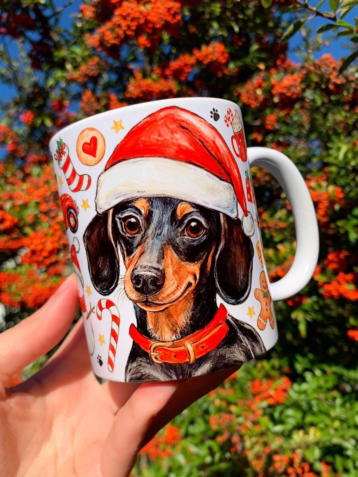 A bögrén cser színű tacskó látható kézzel rajzolt és akvarelles stílusban. A kutyát mézeskalács, cukorpálca és karácsonyi díszek veszik körül. Meleg, ünnepi hangulatú kerámia darab.