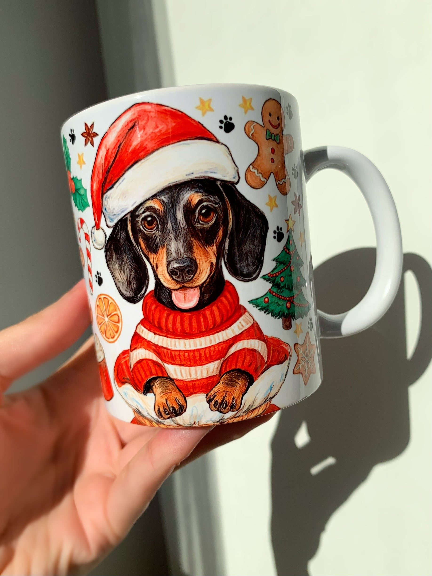 Kézzel festett tacskó mintás karácsonyi kerámia bögre, meleg, természetes akvarellárnyalatokkal. A bájos, részletes kutyaportré ünnepi dekorációkkal egészül ki. Kiváló minőségű, tartós bögrén.