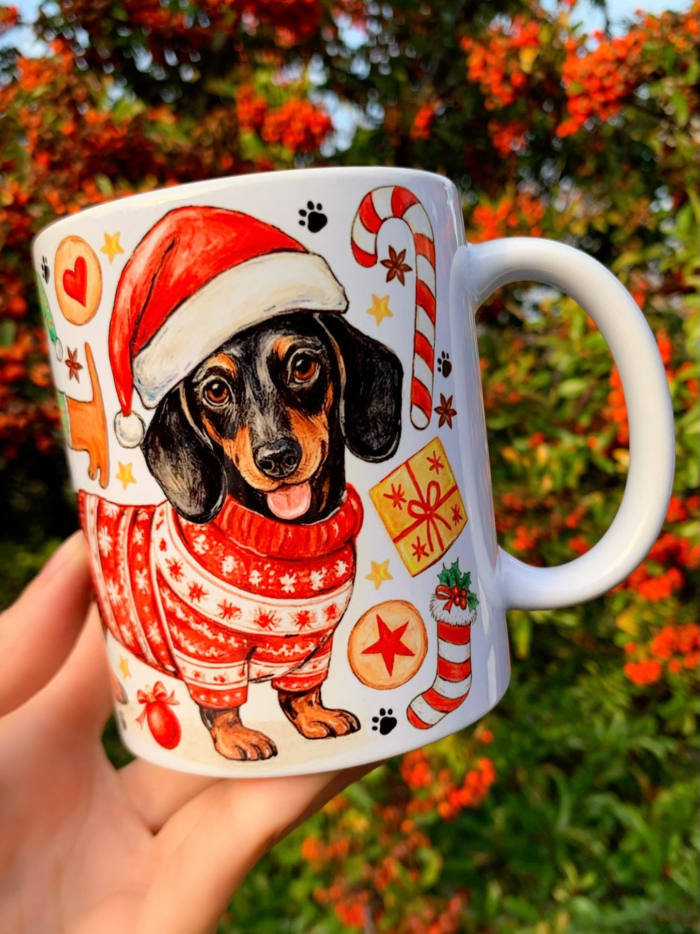 Karácsonyi tacskós bögre kézzel festett akvarell mintával. Álló tacskó piros karácsonyi pulcsiban, körülötte sok apró tacskó Mikulás-sapkában, mézeskalács figurákkal és ünnepi karácsonyi díszekkel. Prémium tacskós ajándék gazdiknak, egyedi kézműves kutyás bögre karácsonyra, iparművészeti illusztrációval.