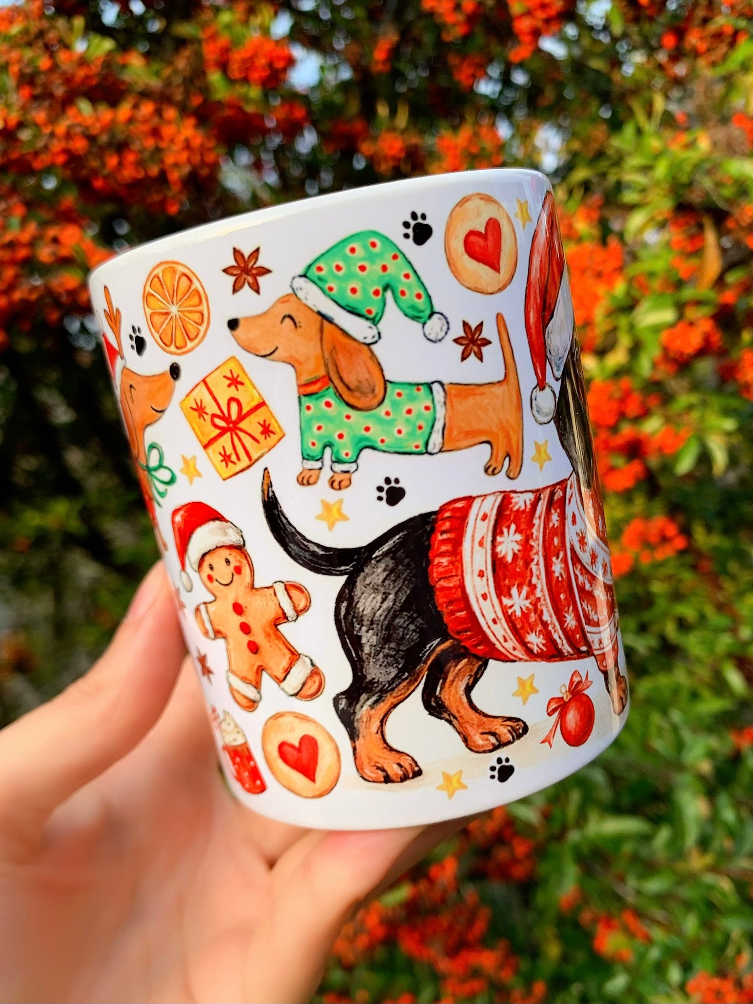 Tacskós bögre karácsonyi mintával, piros és természetes téli árnyalatokkal. A mintán Mikulás-sapkás tacskók és dekoratív karácsonyi elemek szerepelnek. Szerethető ajándék tacskórajongóknak.