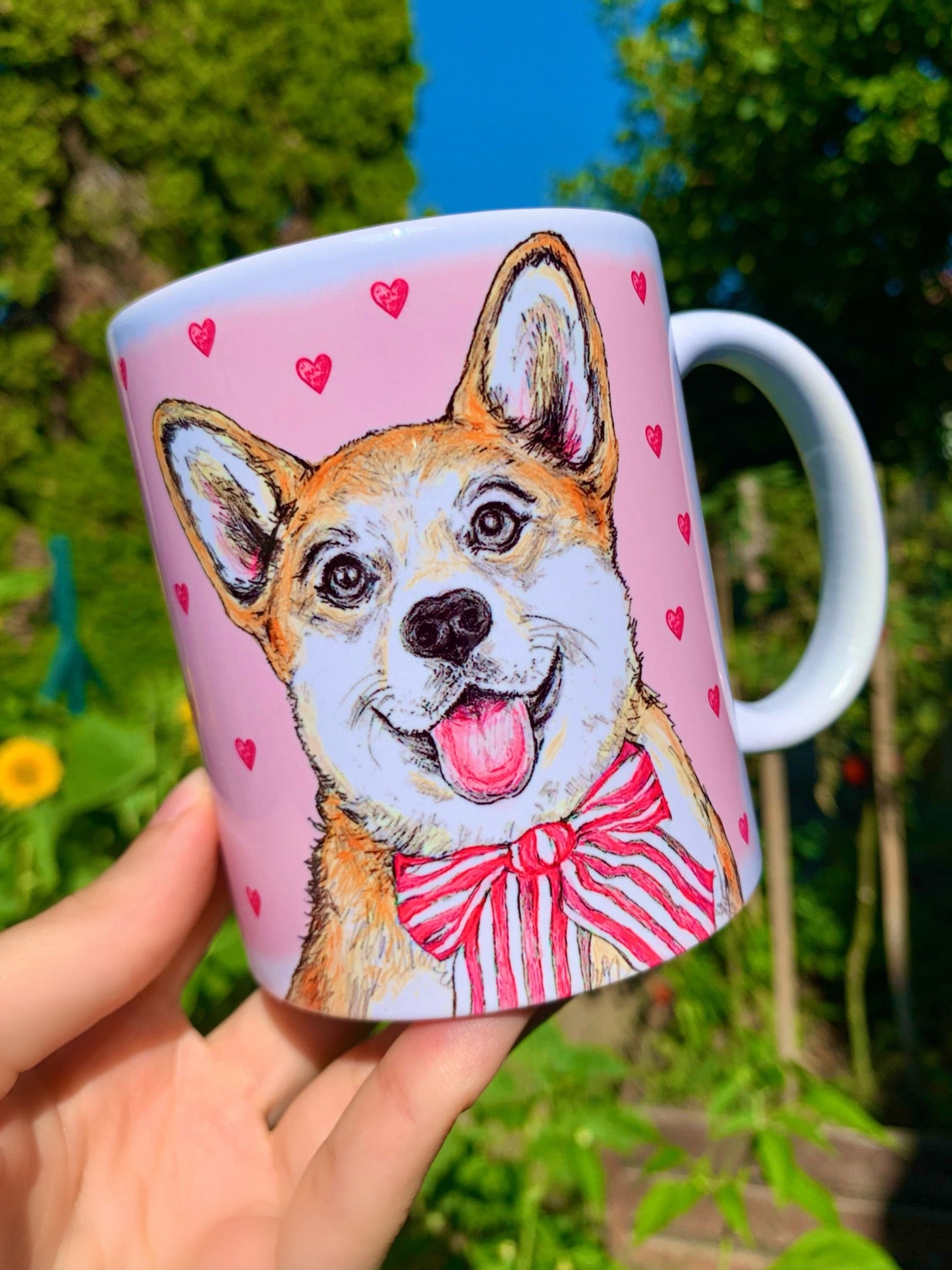 Kerámia bögre kézzel rajzolt, akvarellfestett világosbarna-fehér Corgi kutyával, ülő pózban és rózsaszín szív mintákkal, barátságos mosollyal, fehér háttérrel.