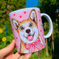 Közelkép a kézzel festett világosbarna-fehér Corgi barátságos arcáról kerámia bögrén, rózsaszín szív mintákkal, fehér háttérrel.