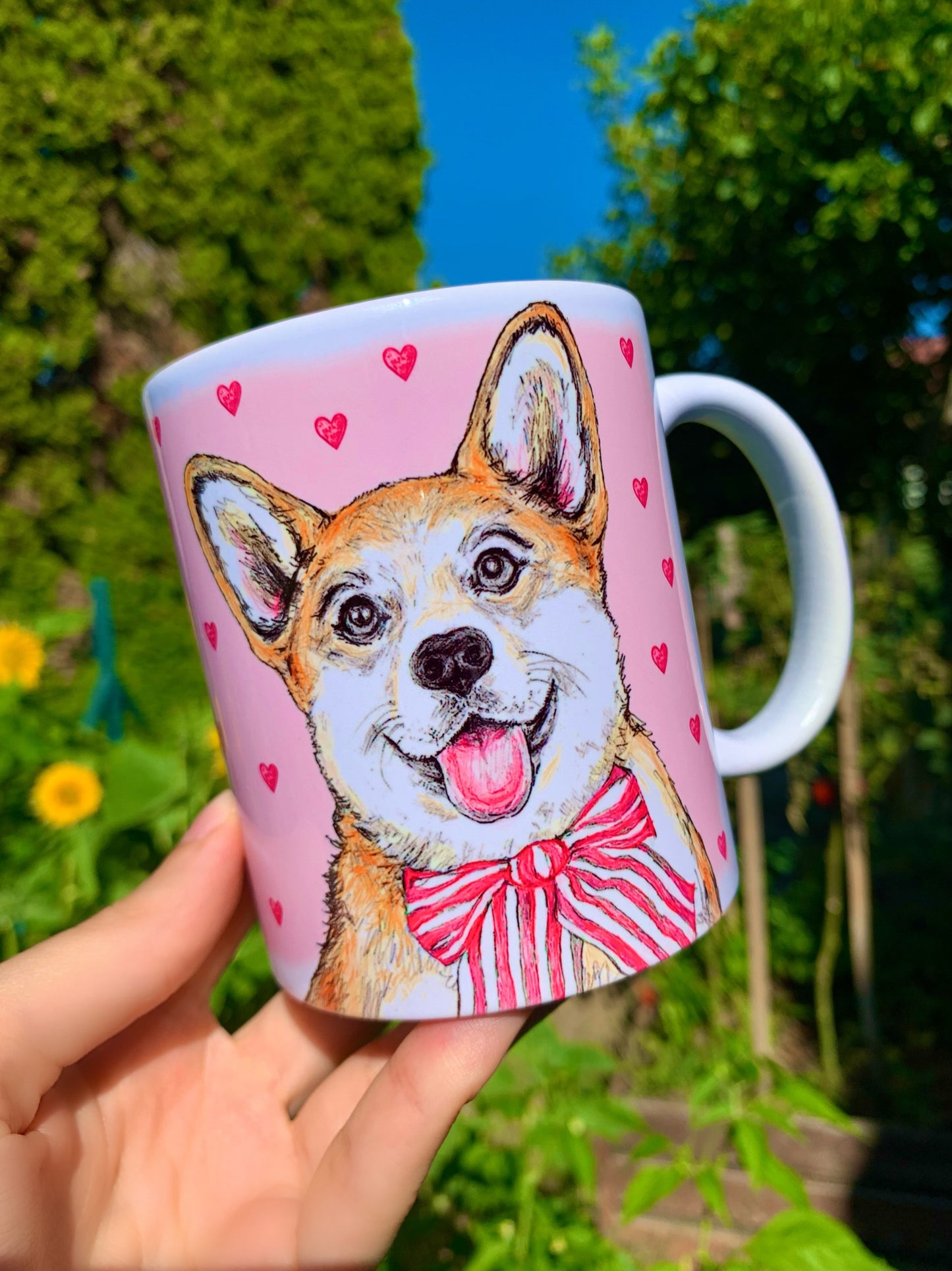 Közelkép a kézzel festett világosbarna-fehér Corgi barátságos arcáról kerámia bögrén, rózsaszín szív mintákkal, fehér háttérrel.