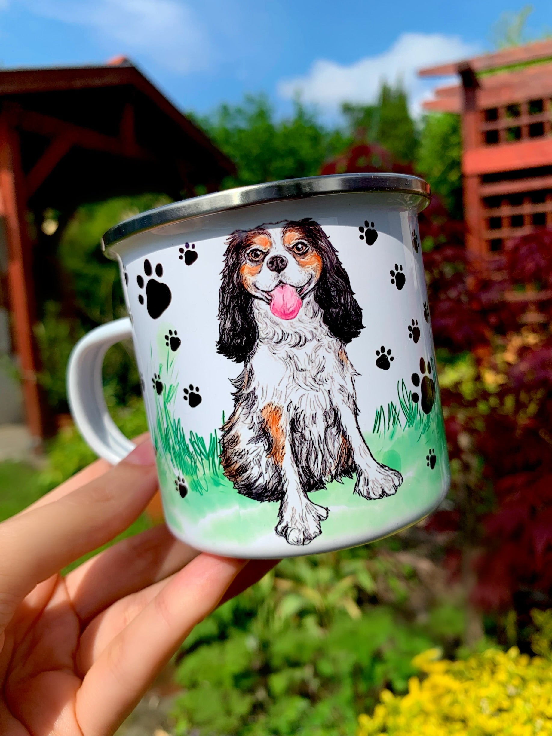 Mosolygó Cavalier spániel illusztráció fém bögrén, barna–fekete–fehér szőrzet, apró fekete mancs mintás fehér design, kézzel rajzolt és festett stílusban