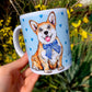 Kerámia bögre kinagyított, mosolygó Corgi fejes illusztrációval és kézzel festett világoskék szív mintákkal, fehér háttérrel.