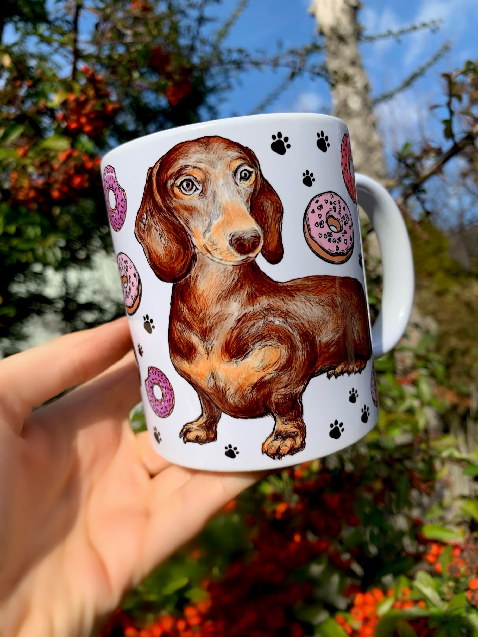 Mosolygó csokibarna tacskó portréja, körülötte színes sütemények és fekete mancsnyomok – kézzel rajzolt és festett kerámia bögrén, ajándéknak is tökéletes.