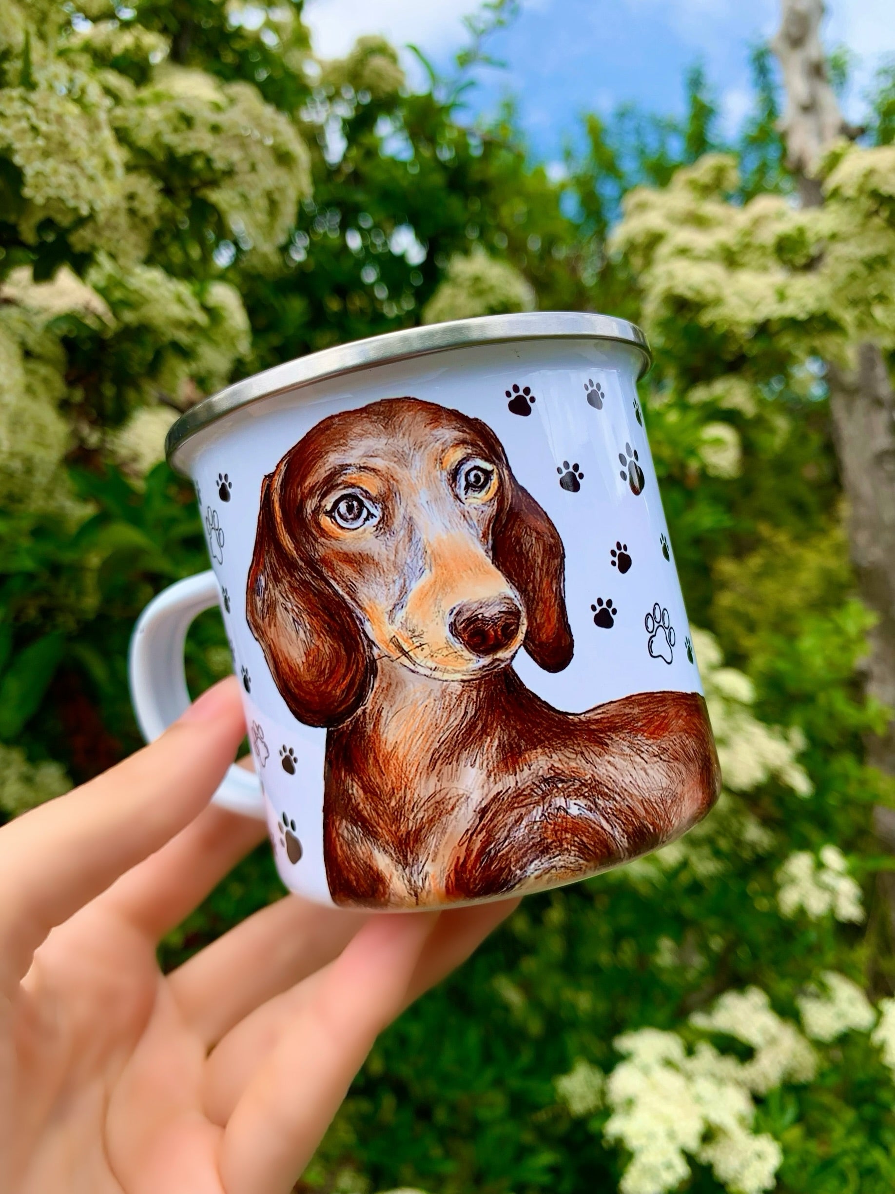 Chocolate tacskó portré akvarell stílusban, lágy ecsetvonásokkal és fényes bundaszínnel, játékos fekete mancsnyom mintával, tartós fém bögrén.